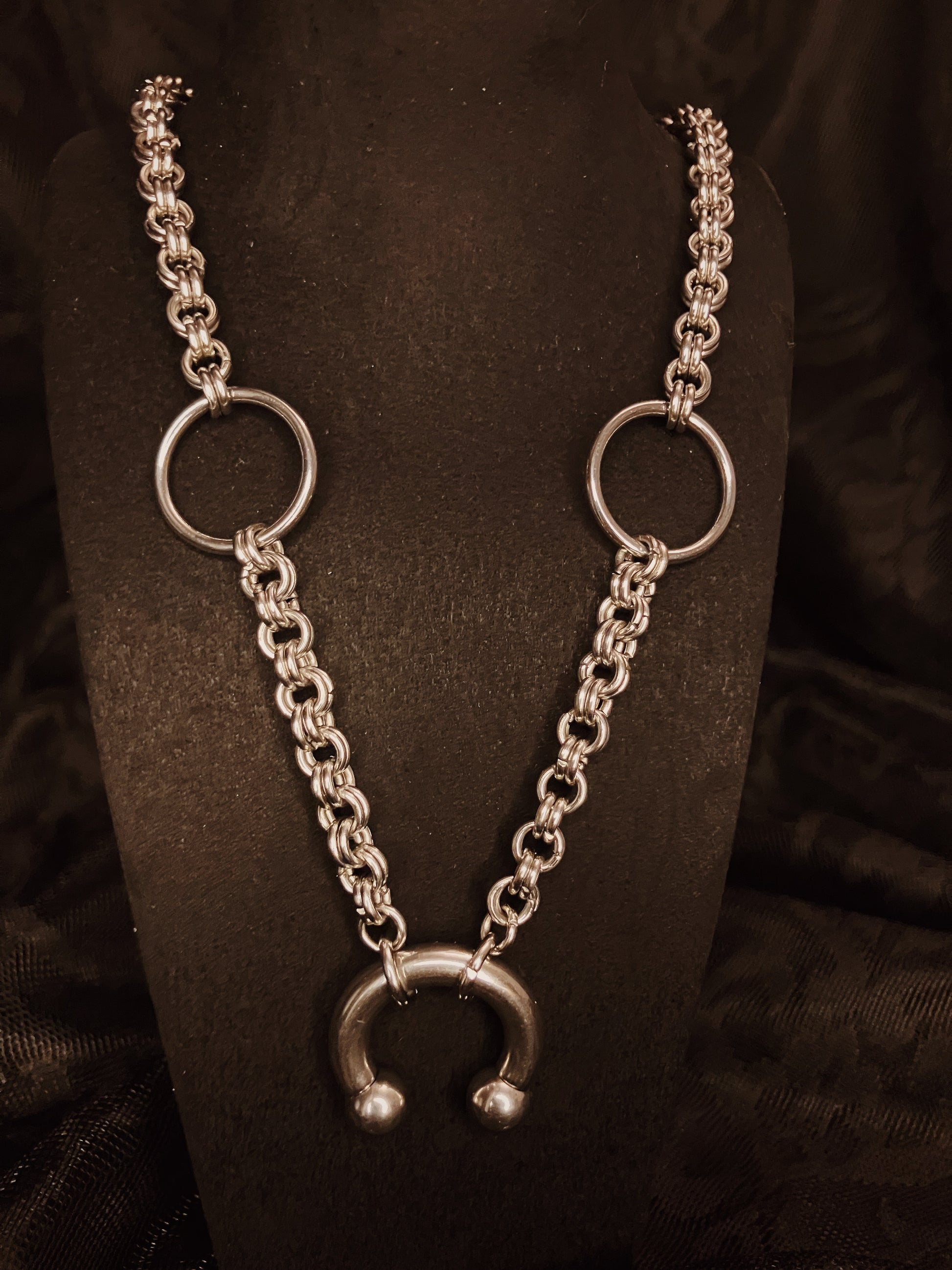 Silver chain necklace with circular and horseshoe-shaped pendants on a black stand.